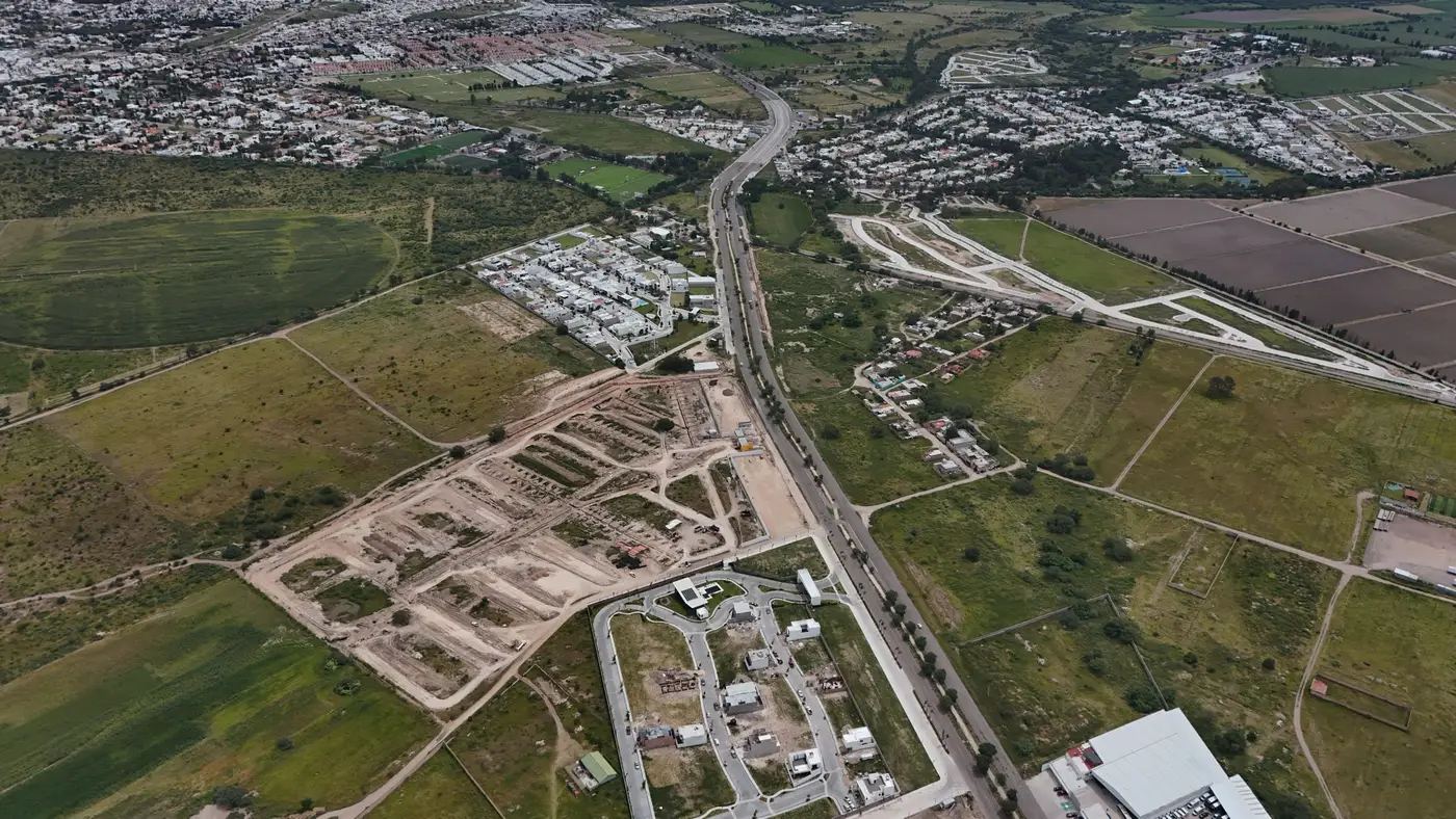 Aerial view of a paved road cutting through farmland toward a new subdivision under construction at a city's edge, with established neighborhoods beyond.